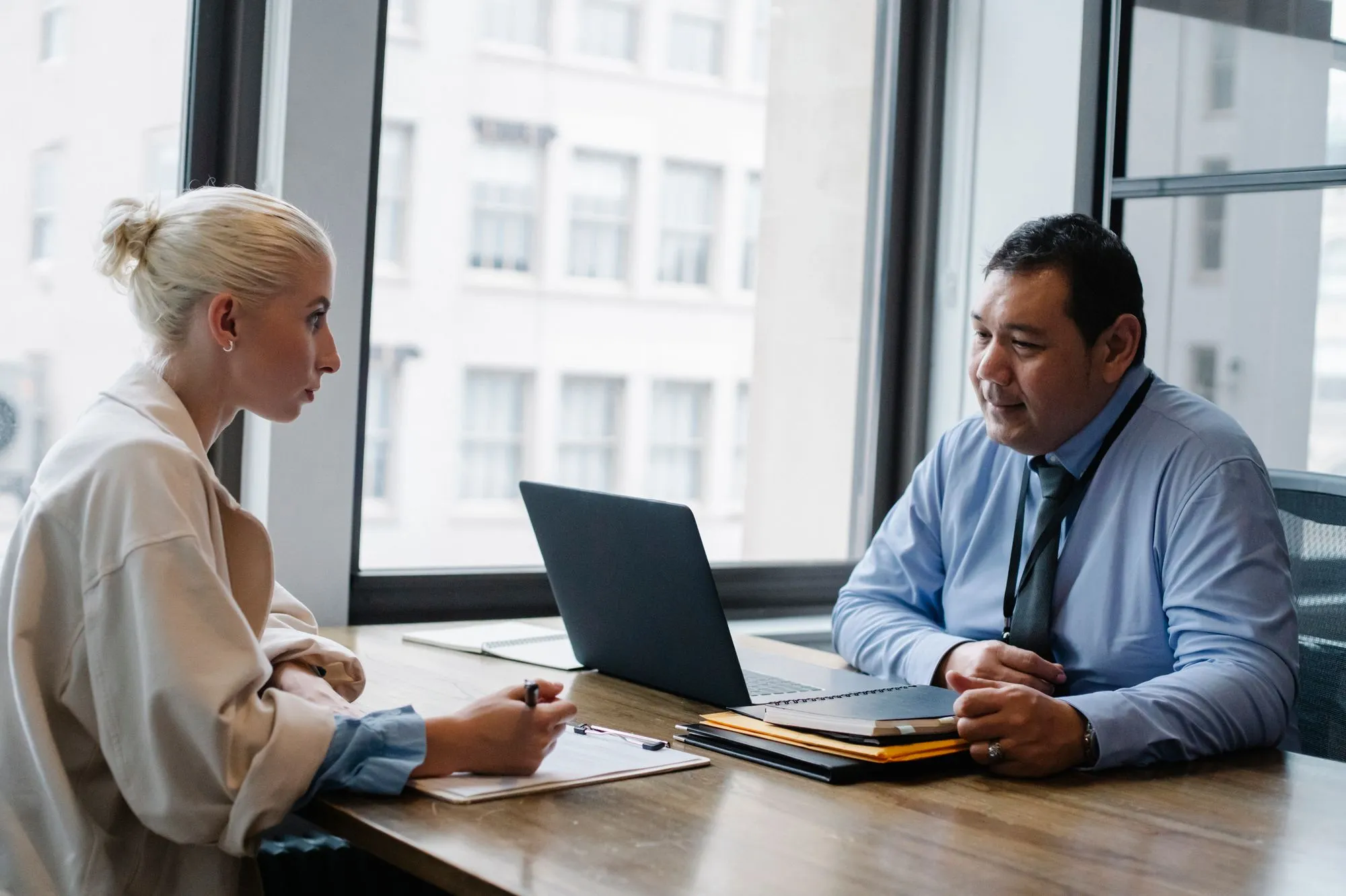A young woman with a clipboard sits across a desk from a smiling man in a shirt and tie during a job interview